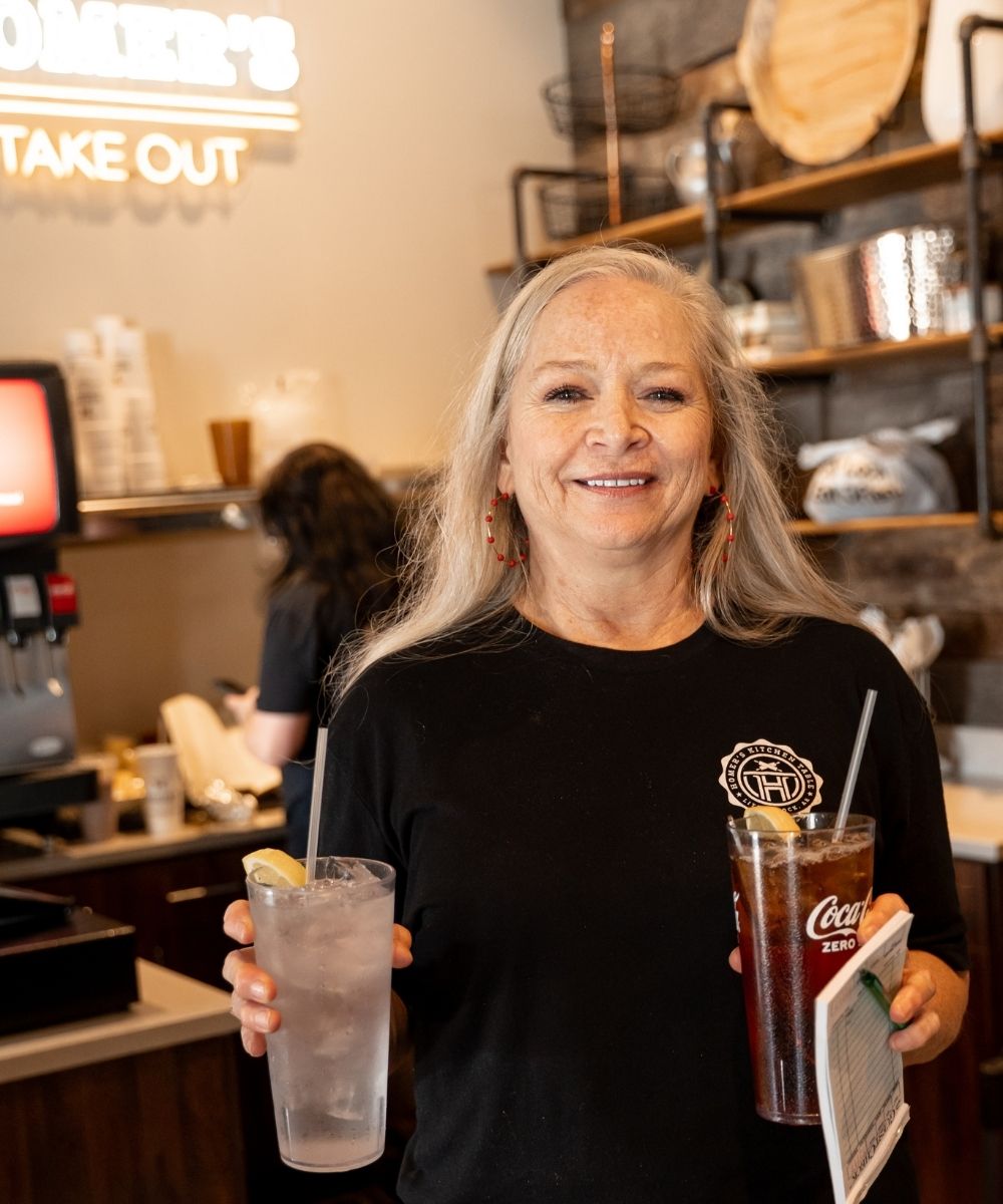 A smiling woman with long gray hair holds a glass of water with lemon and a glass of Coca-Cola Zero inside a restaurant, with shelves and a counter in the background.