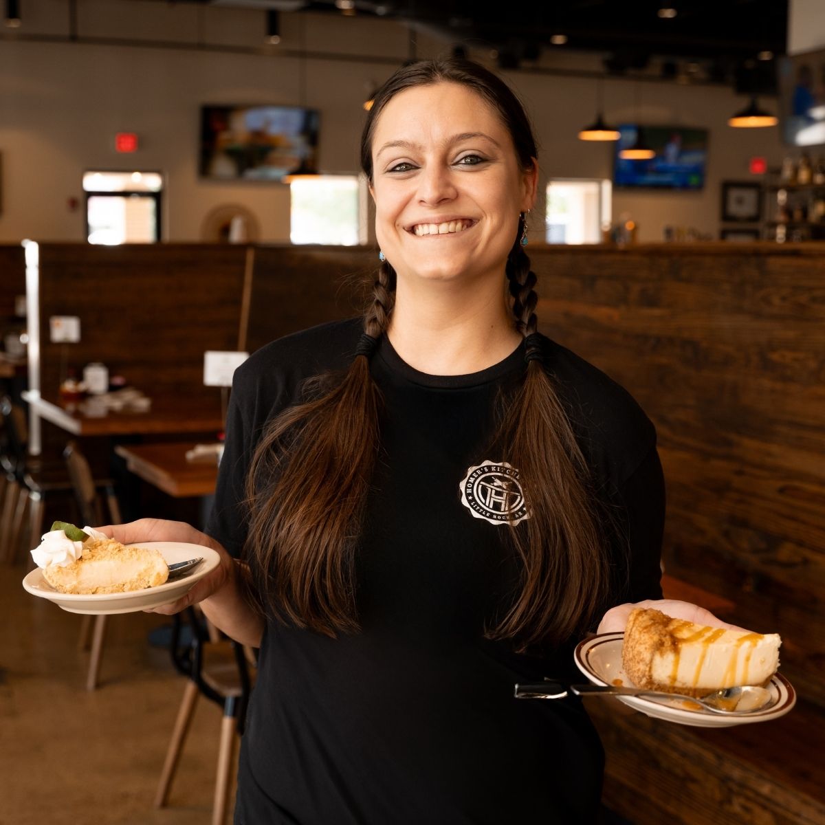 A smiling woman with long brown hair in braids, wearing a black shirt, holds two plates of dessert in a restaurant. The background shows wooden booths and a softly lit, casual dining area.