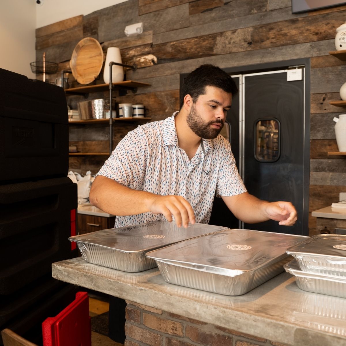 A man in a patterned shirt places lids on aluminum trays of food at a counter in a rustic kitchen or restaurant with wooden walls and shelves holding dishes.