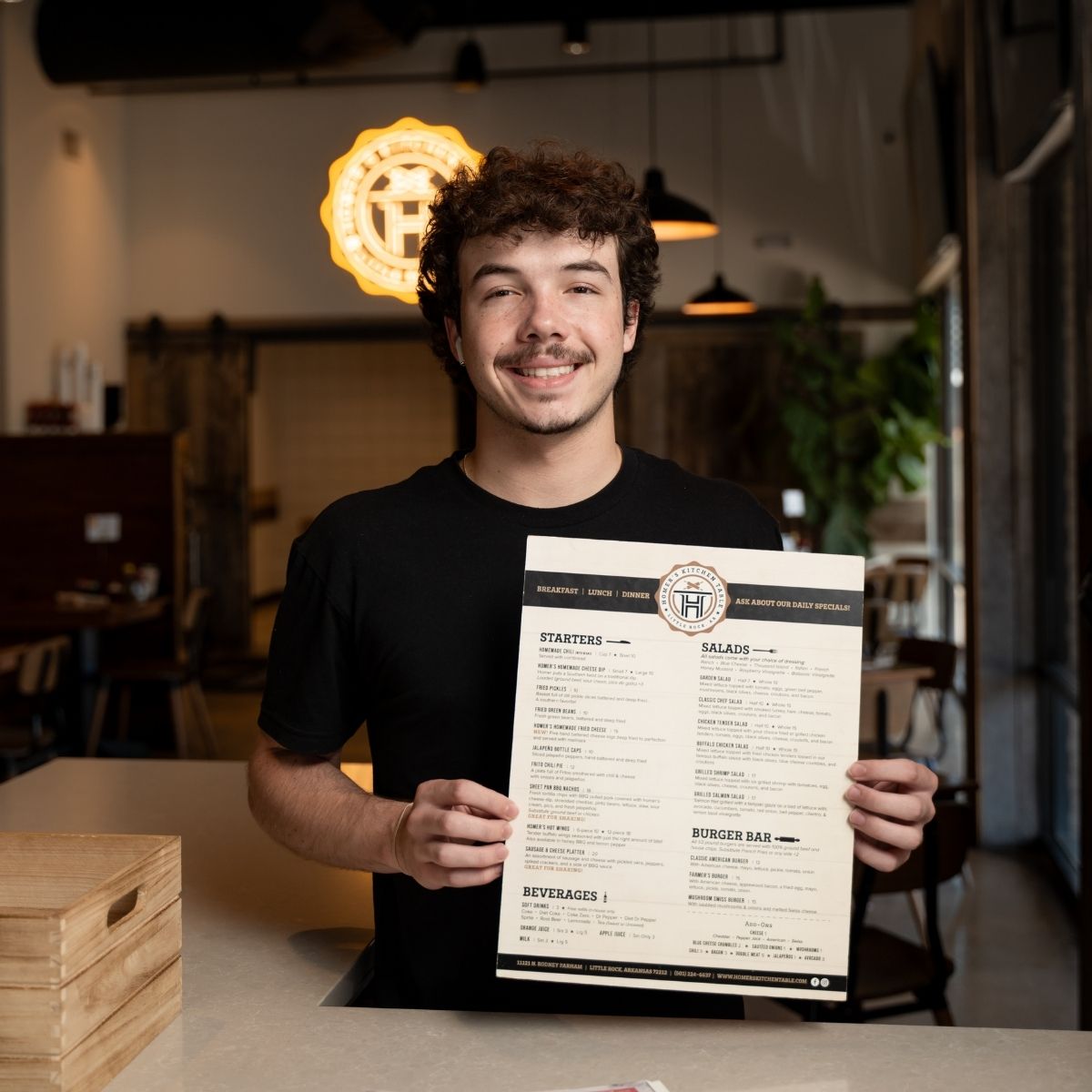 A smiling person with curly hair and a mustache, wearing a black t-shirt, stands behind a counter and holds up a restaurant menu in a warmly lit, modern dining area.