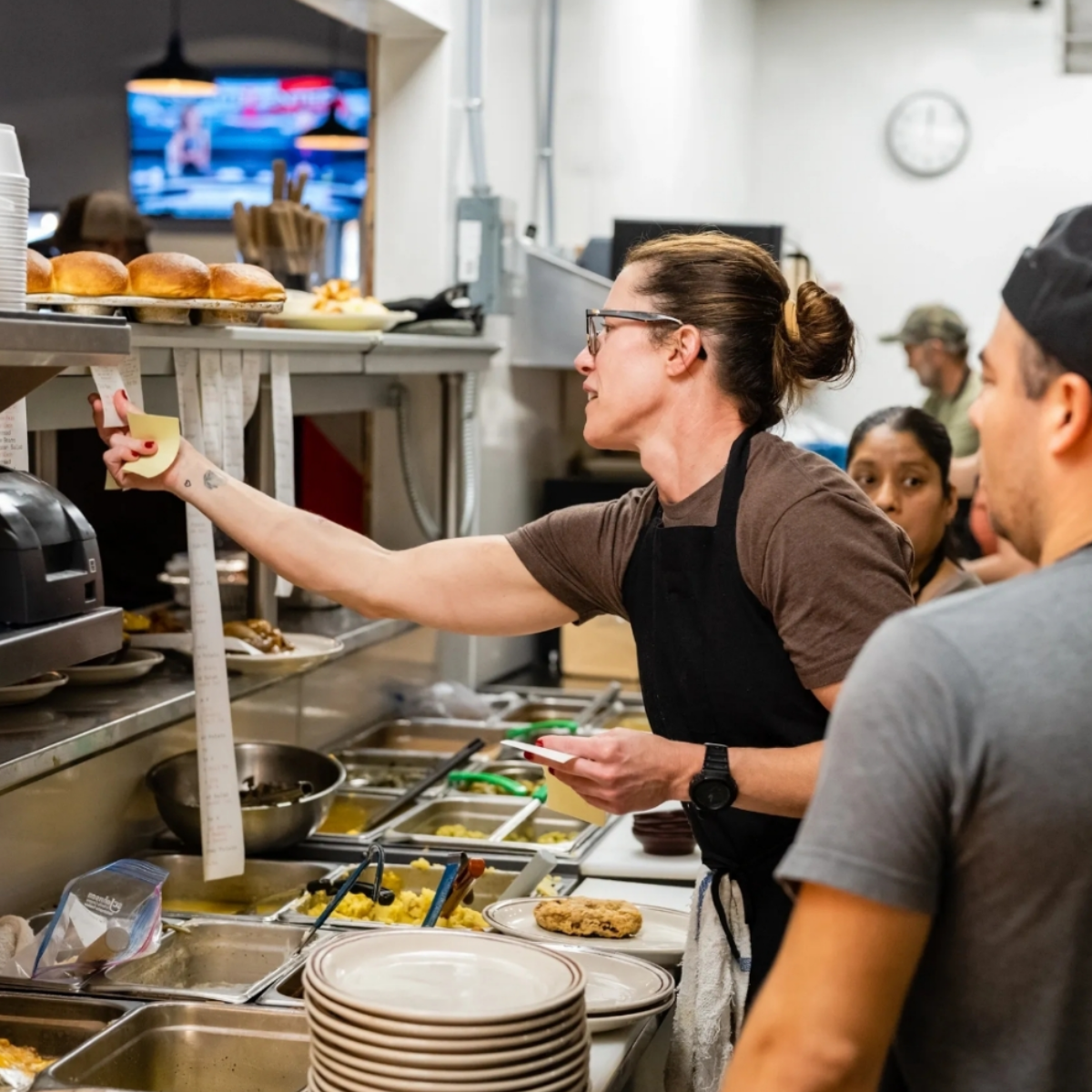 A person wearing glasses and an apron prepares food in a busy restaurant kitchen, reaching for a ticket above a counter lined with plates, ingredients, and cooked burgers. Other staff are visible working nearby.